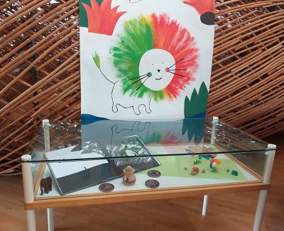 Mise en place sous vitrine de la mini-saynete et d'une partie du jeu "La crinière de Petit lion" sur fond de la bannière de l'exposition.